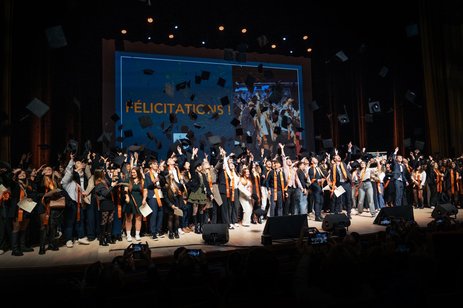 Les diplômés lancent leurs toques sur la scène du Grand Auditorium, devant l’écran « Félicitations ! »